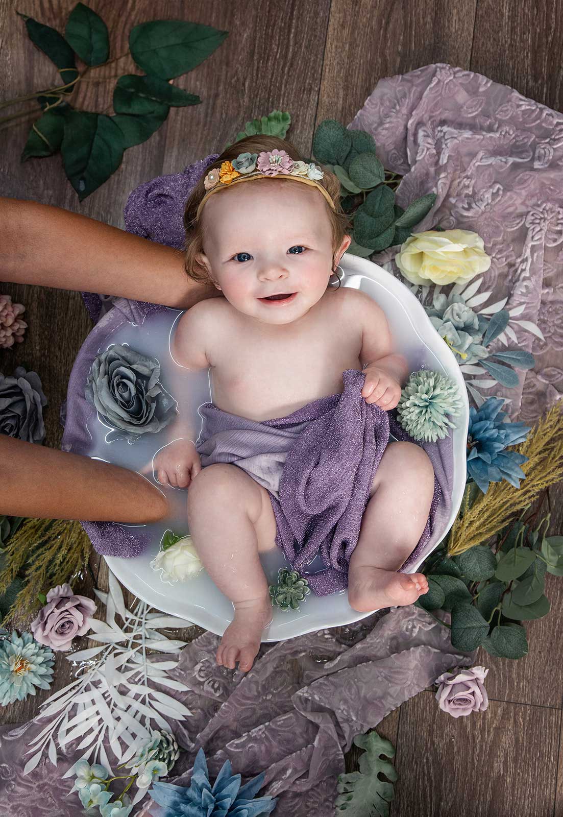 Smiling baby with a flower crown sitting in a milk bath while being held