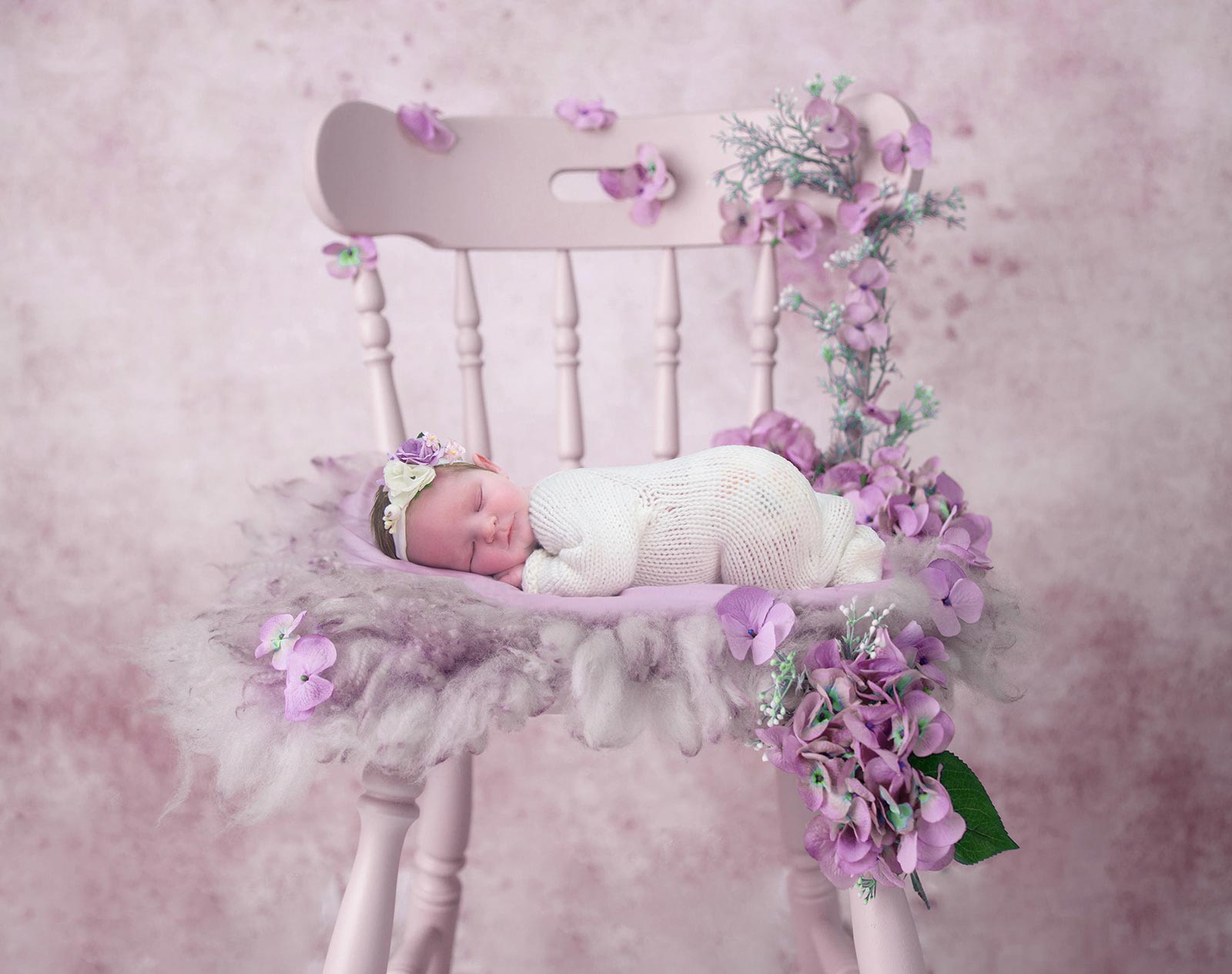 Newborn baby sleeping peacefully on a pink chair, surrounded by purple and pink flowers
