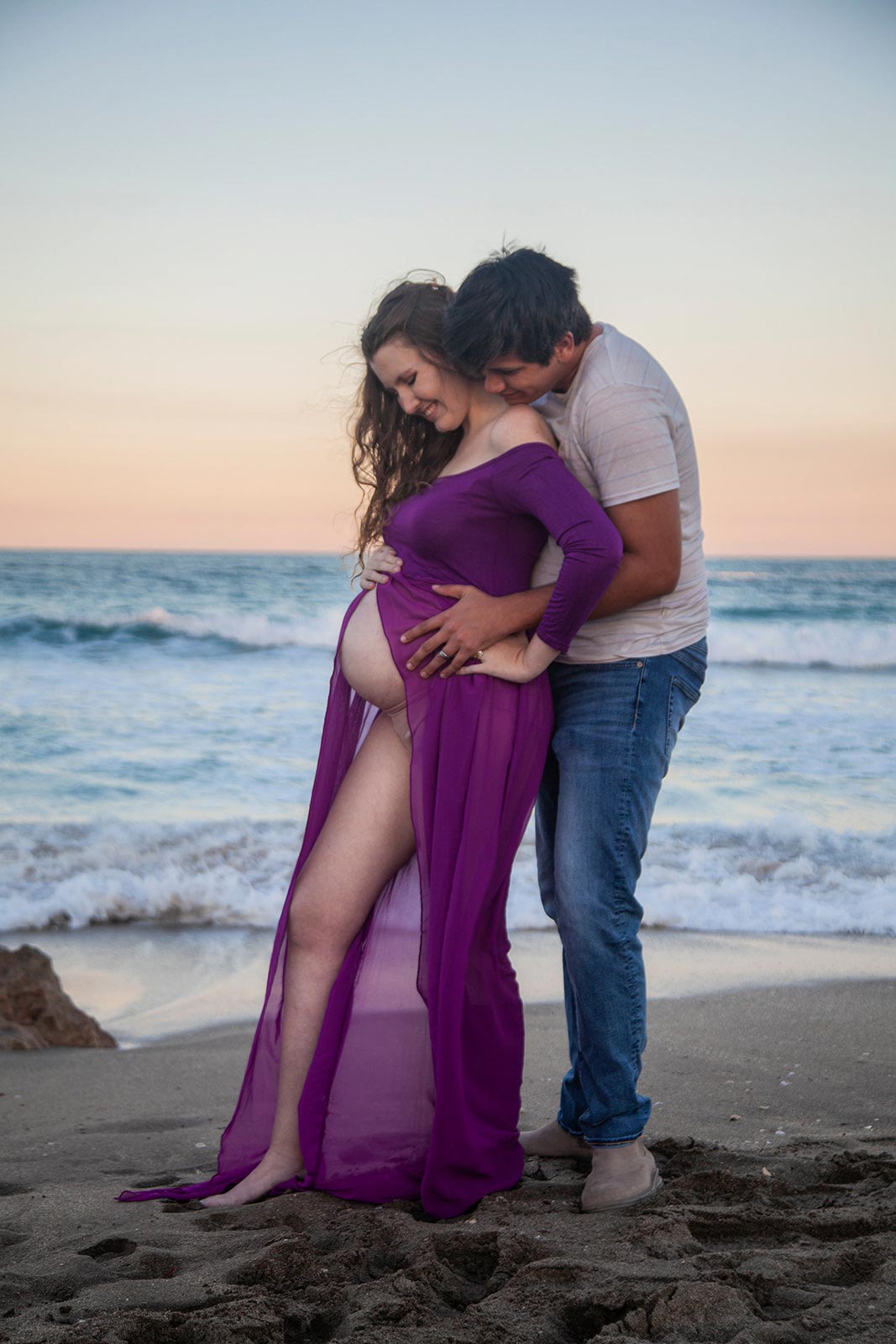 Man standing behind pregnant wife and holding her on the beach