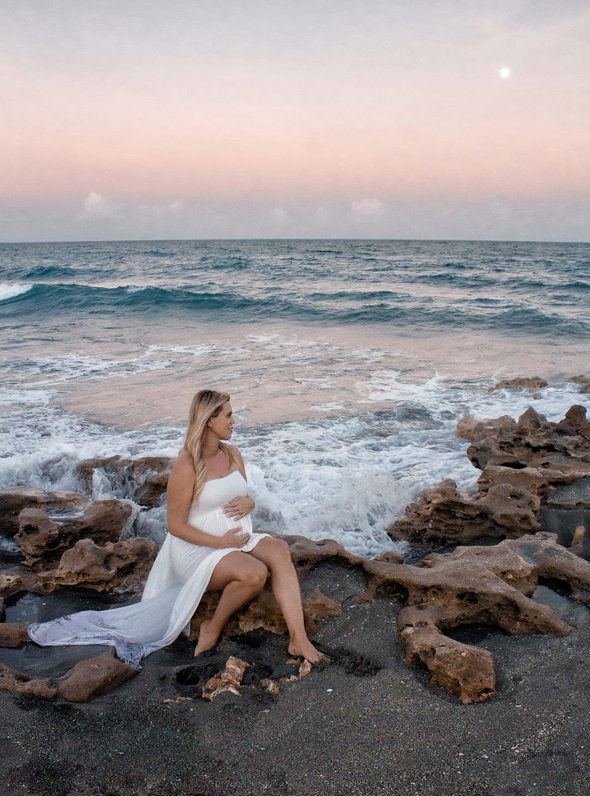 Pregnant woman holding her belly while sitting on rocks by the ocean