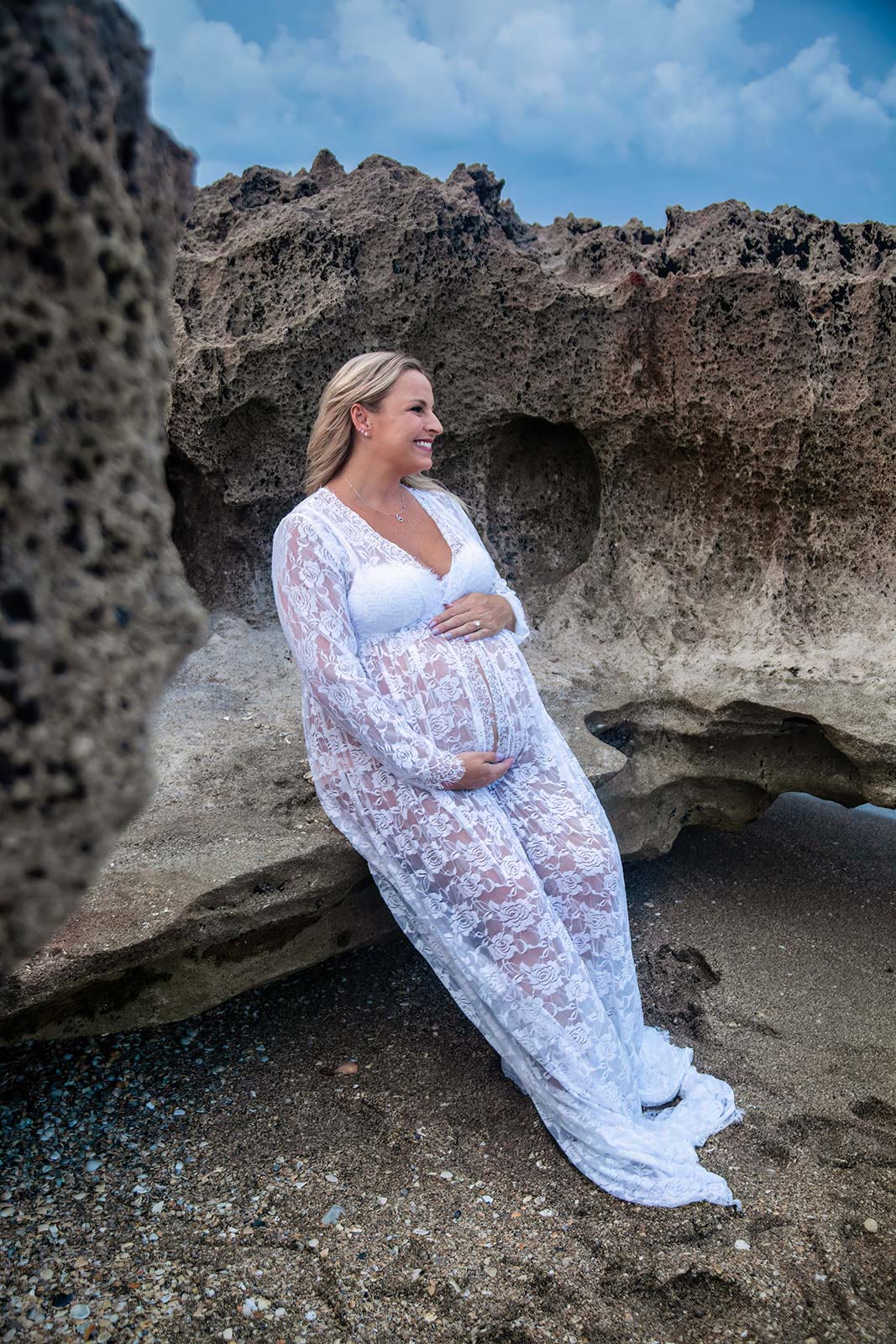 Pregnant woman adorned in white leaning rocks by the beach