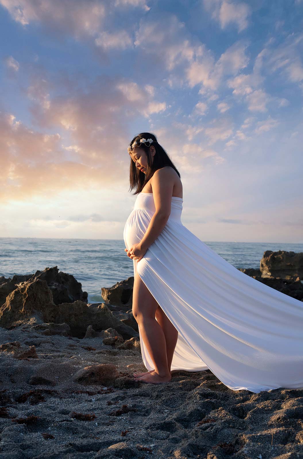 Pregnant woman in white dress standing on rocky beach while the sun sets
