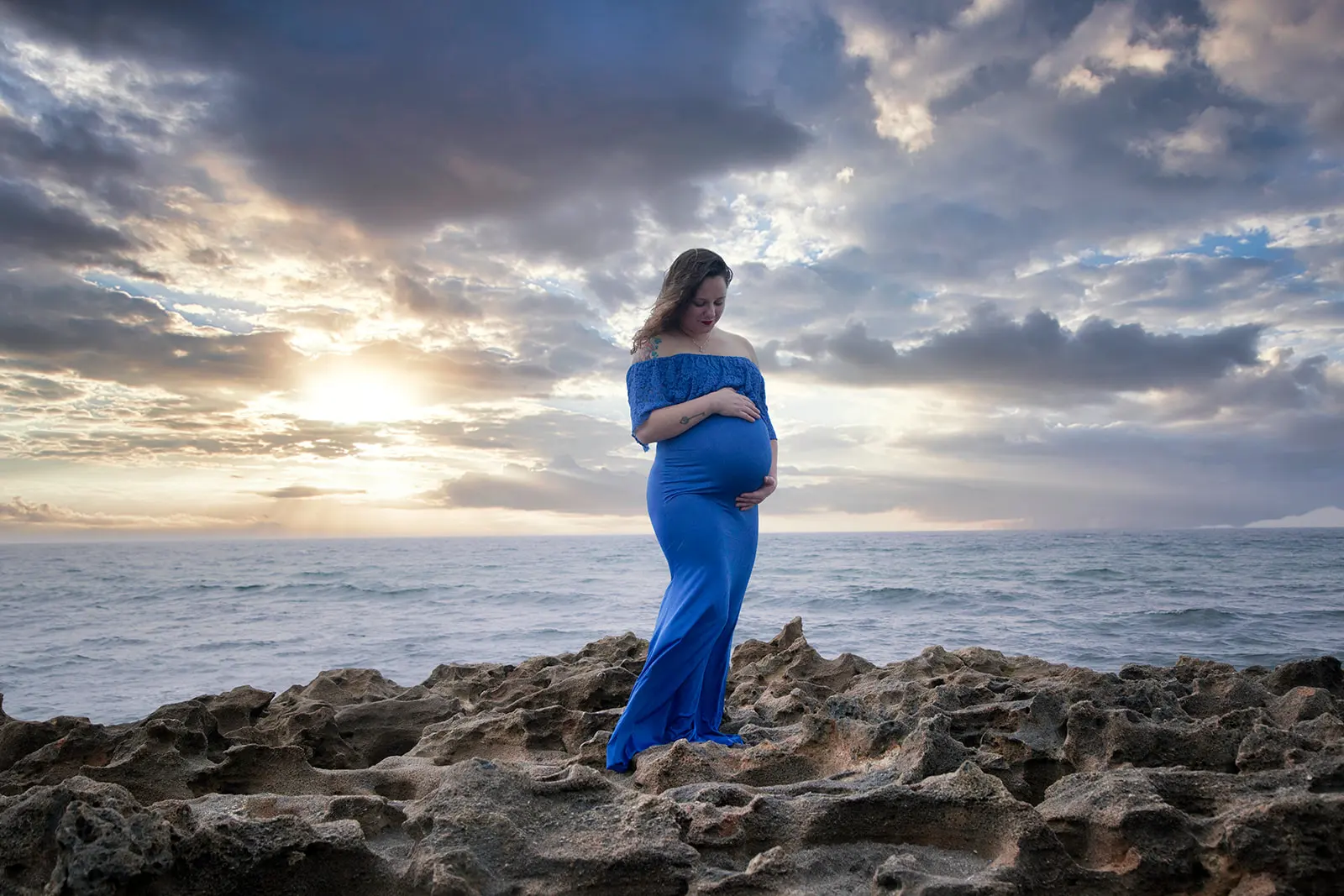 Pregnant woman in blue dress standing on rocks with oceanic background, captured by Nicole Marie Photography in Port St. Lucie