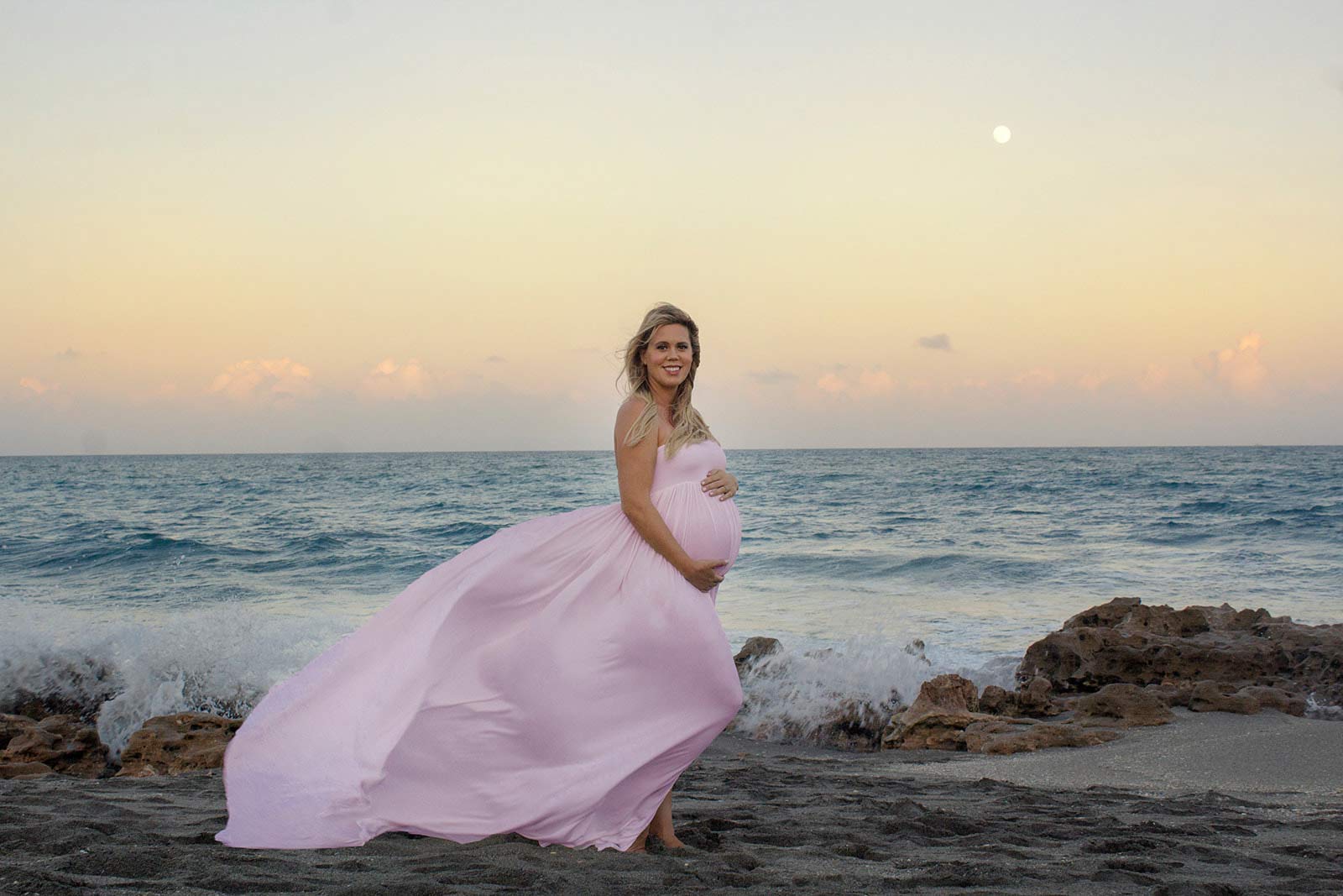 Pregnant woman in pink dress holding her belly by the ocean